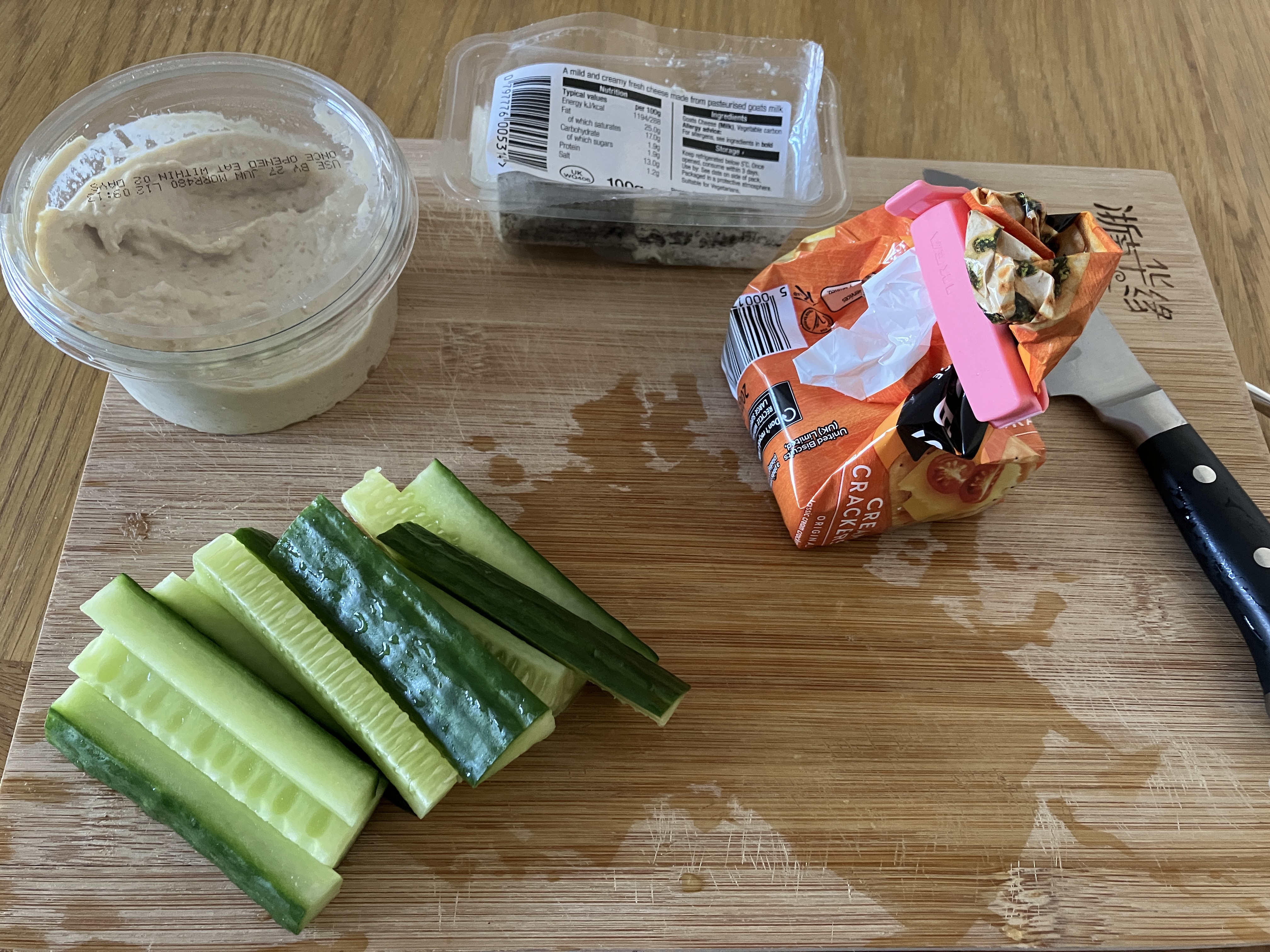 some food on a wooden chopping board: cucumber sticks, a pot of hummus, a pot of goats cheese, and a small pack of jacob's cream crackers